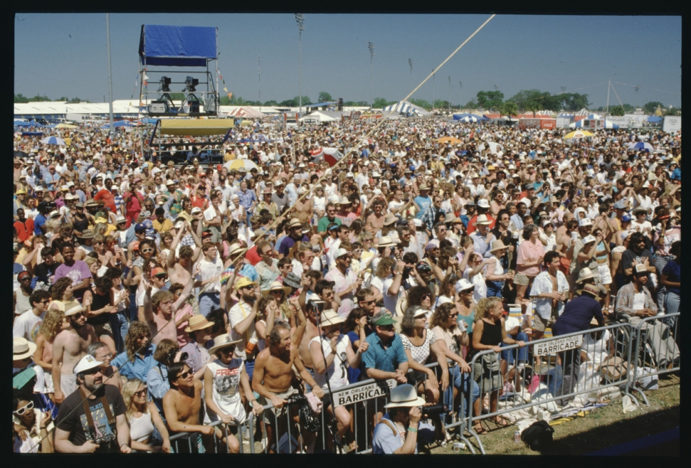 Large outdoor concert audience packed closely together, many are wearing hats and sunglasses. There are sections marked by barricades, and a tower structure with people observing the event. Tents and trees are visible in the background.