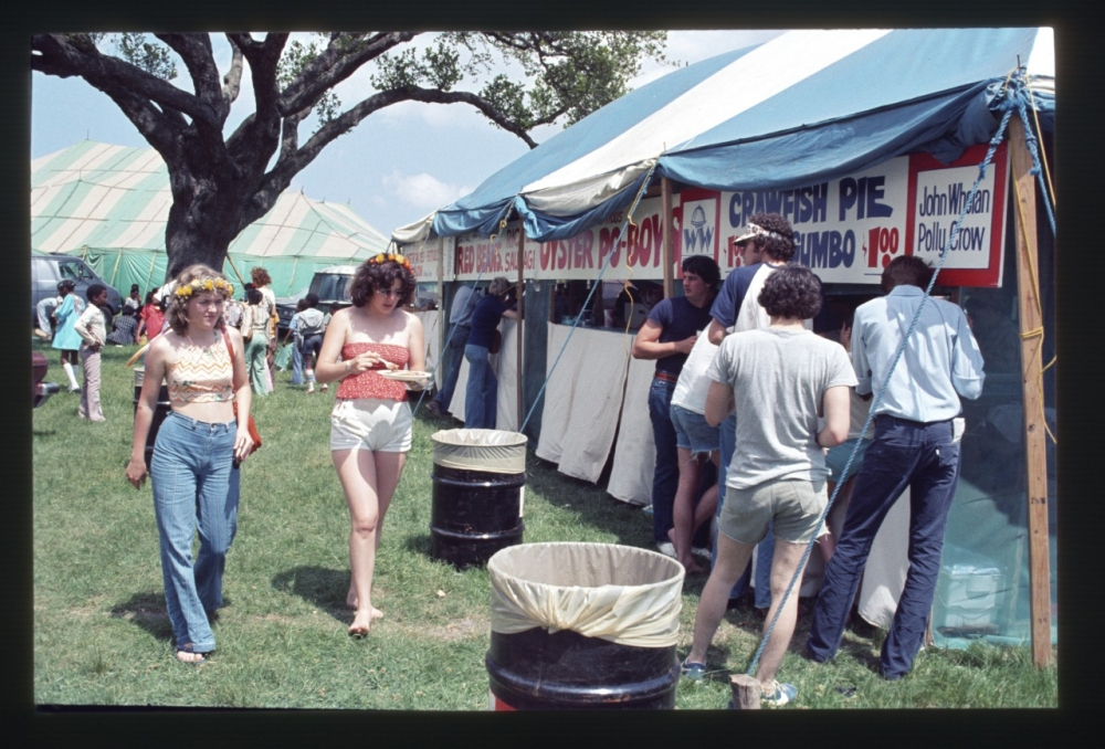 People walk near a food tent at an outdoor festival. The tent offers dishes like crawfish pie and gumbo. Signs and trees are visible, and attendees wear casual summer clothing, some with flower headbands. A large tree stands in the background.