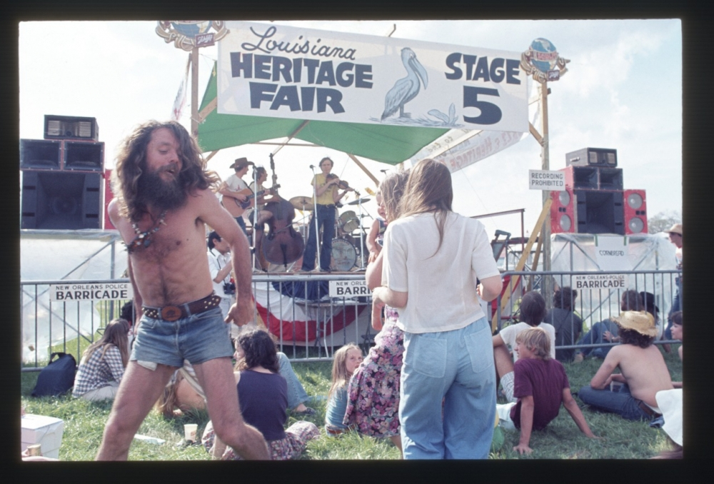 A bearded man dances joyfully at the Louisiana Heritage Fair Stage 5, while others sit on the grass and watch a band perform in the background. The atmosphere is lively, and a banner with a pelican is visible above the stage.