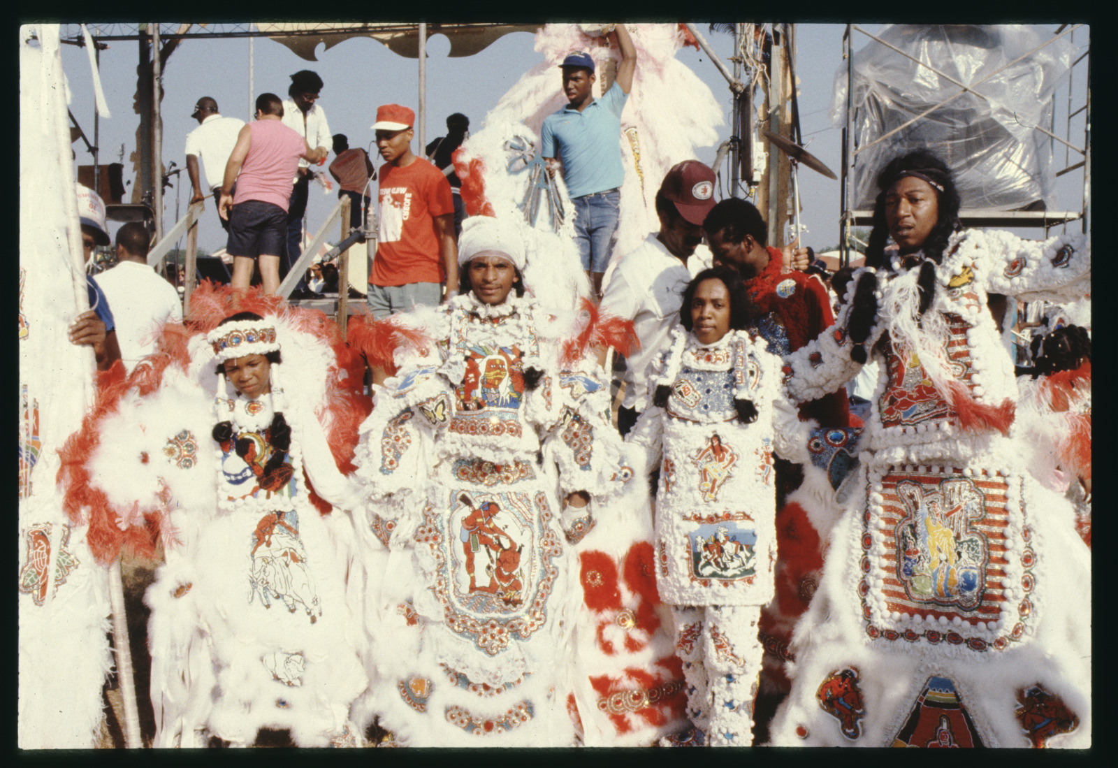 A group of people dressed in vibrant, ornate costumes with feathers and intricate beadwork stand together. The costumes feature red, white, and colorful designs. There are people in the background on a stage or platform.