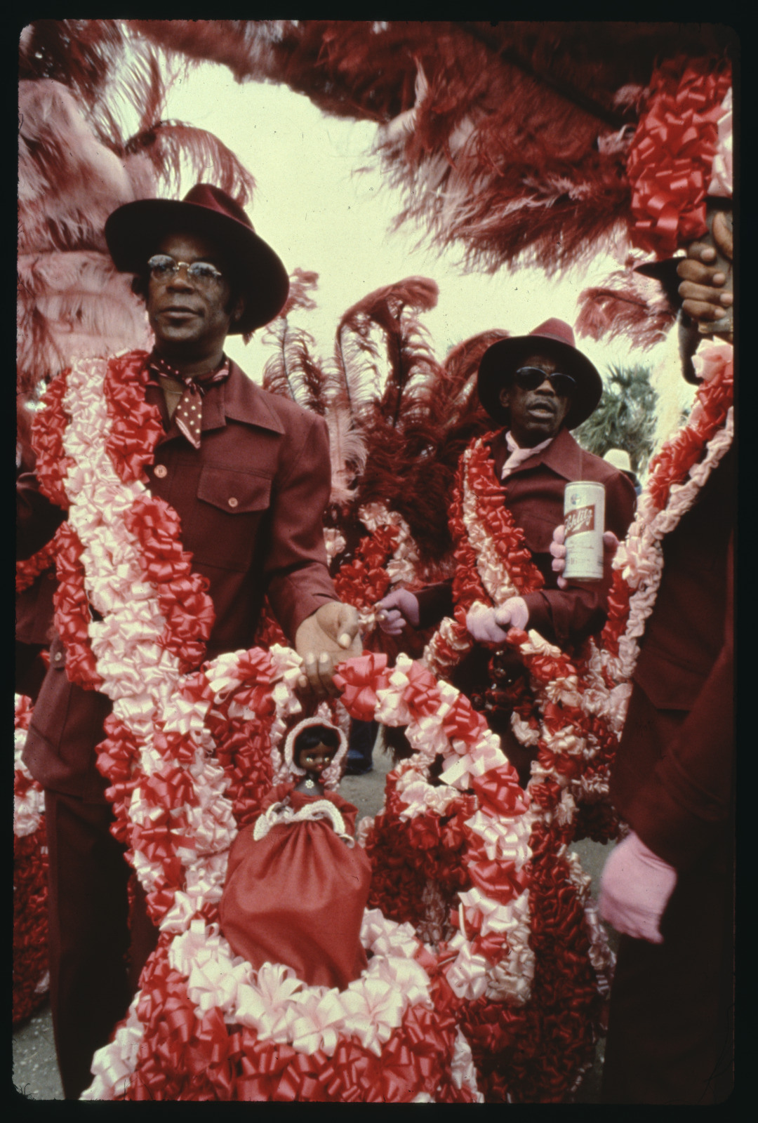 Two men dressed in maroon suits and hats, adorned with bright red and white feathered sashes, participate in a festive parade. One pushes a stroller decorated with matching colors, containing a doll. Both are surrounded by vibrant feathered decorations.
