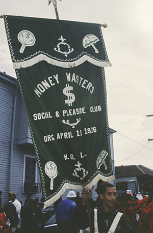 A parade participant carries a green banner reading Money Wasters Social & Pleasure Club, Org. April 11, 1976, N.O. LA adorned with dollar signs and fan motifs. A group of people and houses are visible in the background.