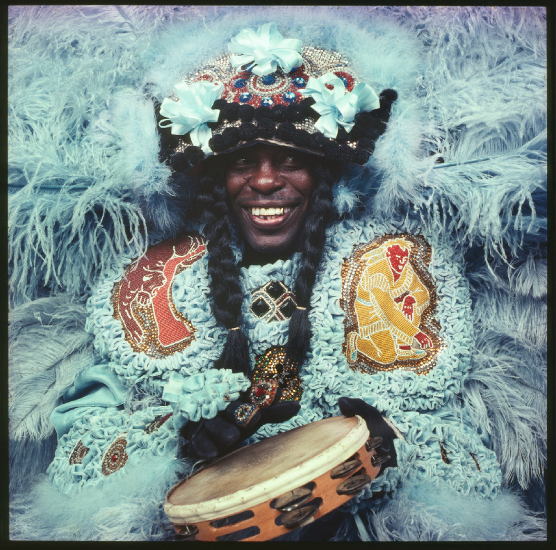 A person wearing an elaborate, colorful costume adorned with feathers, beads, and sequins, smiles while holding a tambourine. The costume features intricate designs and floral elements in shades of blue, red, and gold.
