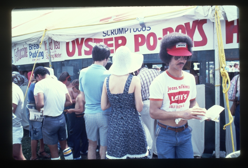 People are lined up at a food stall selling oyster po boys. A man in a red visor and graphic T-shirt is reading a pamphlet, while a woman in a white hat stands nearby. The atmosphere is casual and crowded.