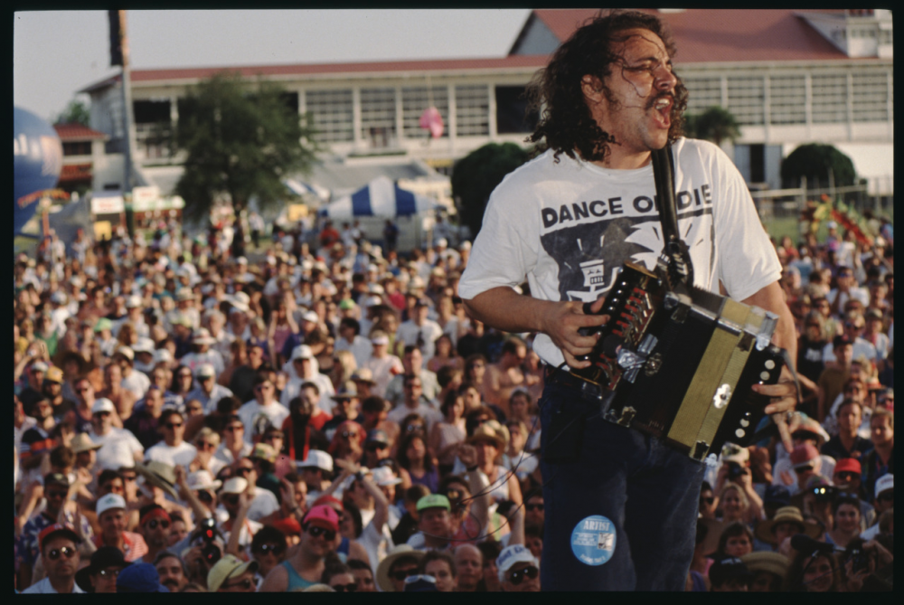 Musician passionately playing an accordion on stage, wearing a Dance or Die shirt. A large, lively crowd is gathered in an outdoor venue, with tents and a building in the background under a clear sky.