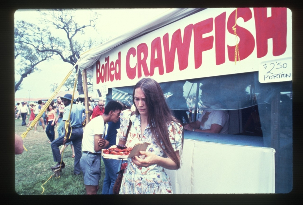 A young woman with long hair holds a tray of crawfish in front of a booth with a sign reading Boiled CRAWFISH at a festival. People are gathered around, enjoying the outdoor event. Trees are visible in the background.