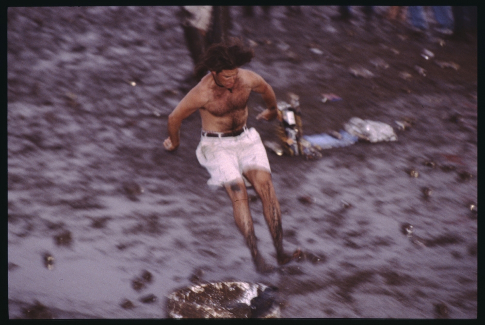 A shirtless person with long hair, wearing white shorts, runs energetically through a muddy field. Trash is scattered around, creating a chaotic environment. The person appears carefree and lively amidst the messy surroundings.