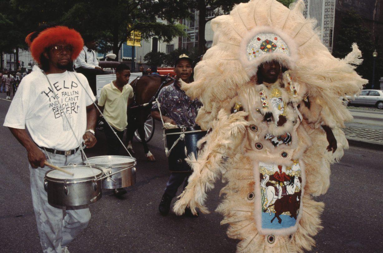 A man in a large, feathered costume with intricate designs walks in a parade. Beside him, another man plays a drum, wearing a red hat and a shirt that reads HELP THE FALLEN AND I CANT REACH MY BEER. People and buildings are visible in the background.