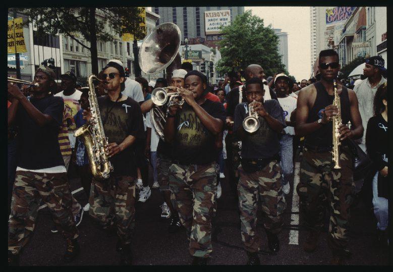 A marching band plays instruments including saxophones, trumpet, and sousaphone during a street parade. The musicians wear matching outfits with dark shirts and camouflage pants. A crowd watches in the background.