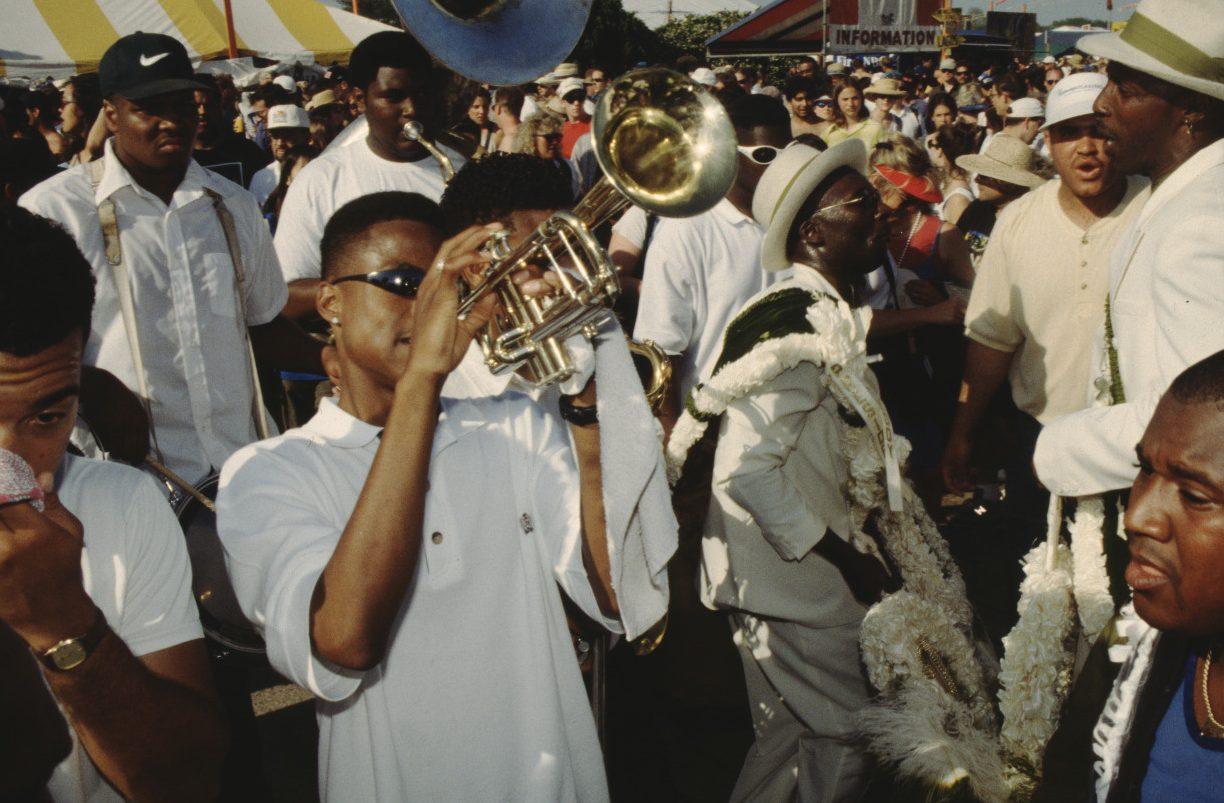 A marching band performs at a lively outdoor event. Musicians play brass instruments, dressed in white. Nearby, a man in a decorated suit and hat carries a parasol adorned with flowers. The crowd watches enthusiastically under colorful tents.