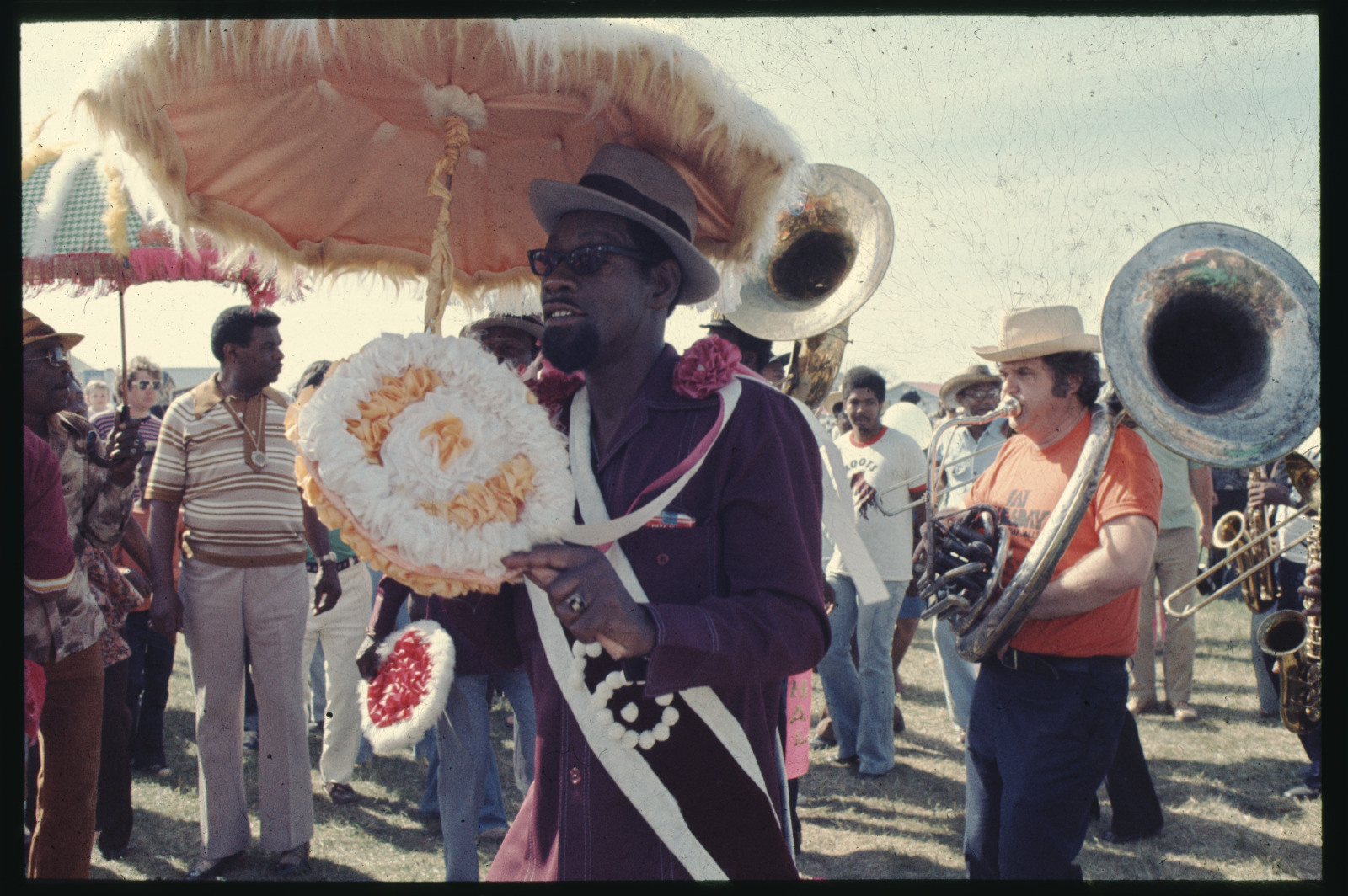 A festive parade with a man in a maroon outfit holding a decorative wreath and standing under a large orange parasol. Musicians, including horn players, accompany him. Onlookers and participants surround the scene in a sunny outdoor setting.