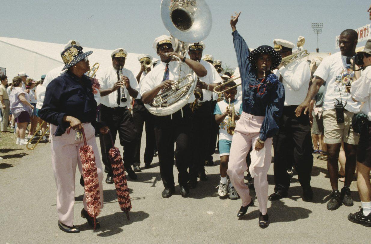 A lively marching band with musicians playing brass instruments parades down a sunny street. A woman in the foreground is energetically dancing, while spectators watch and cheer. Everyone is dressed in bright, festive clothing.