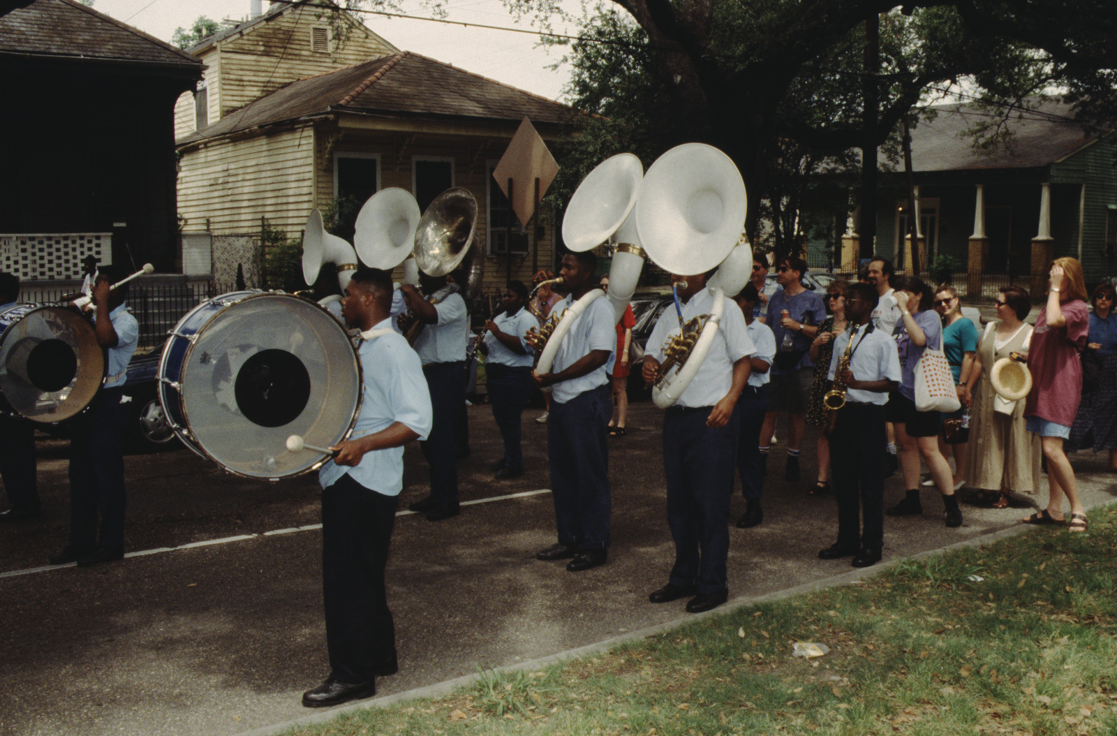 A marching band parades down a street, with musicians playing sousaphones, saxophones, and drums. The band members wear light blue shirts and dark pants. Onlookers watch from the sidewalk, and houses are visible in the background.