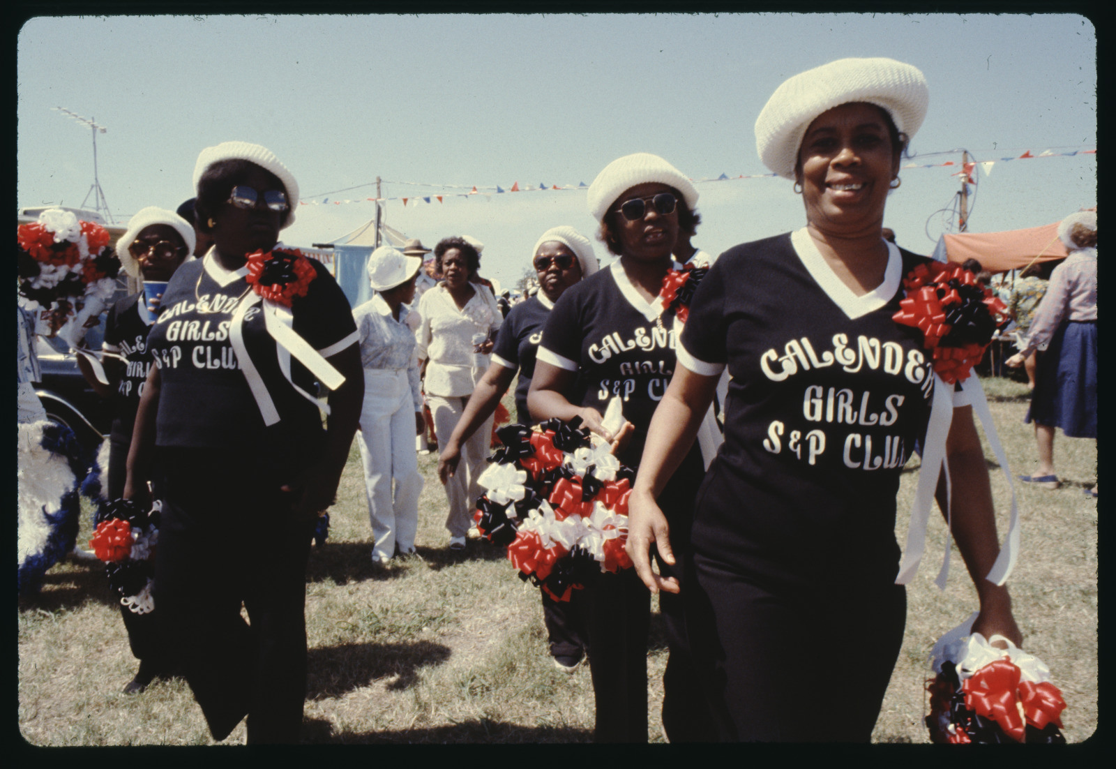 A group of people wearing matching Calender [sic] Girls Sap Club outfits and white hats walk together outdoors. They are holding red and white pom-poms, and the background shows tents and festive decorations.