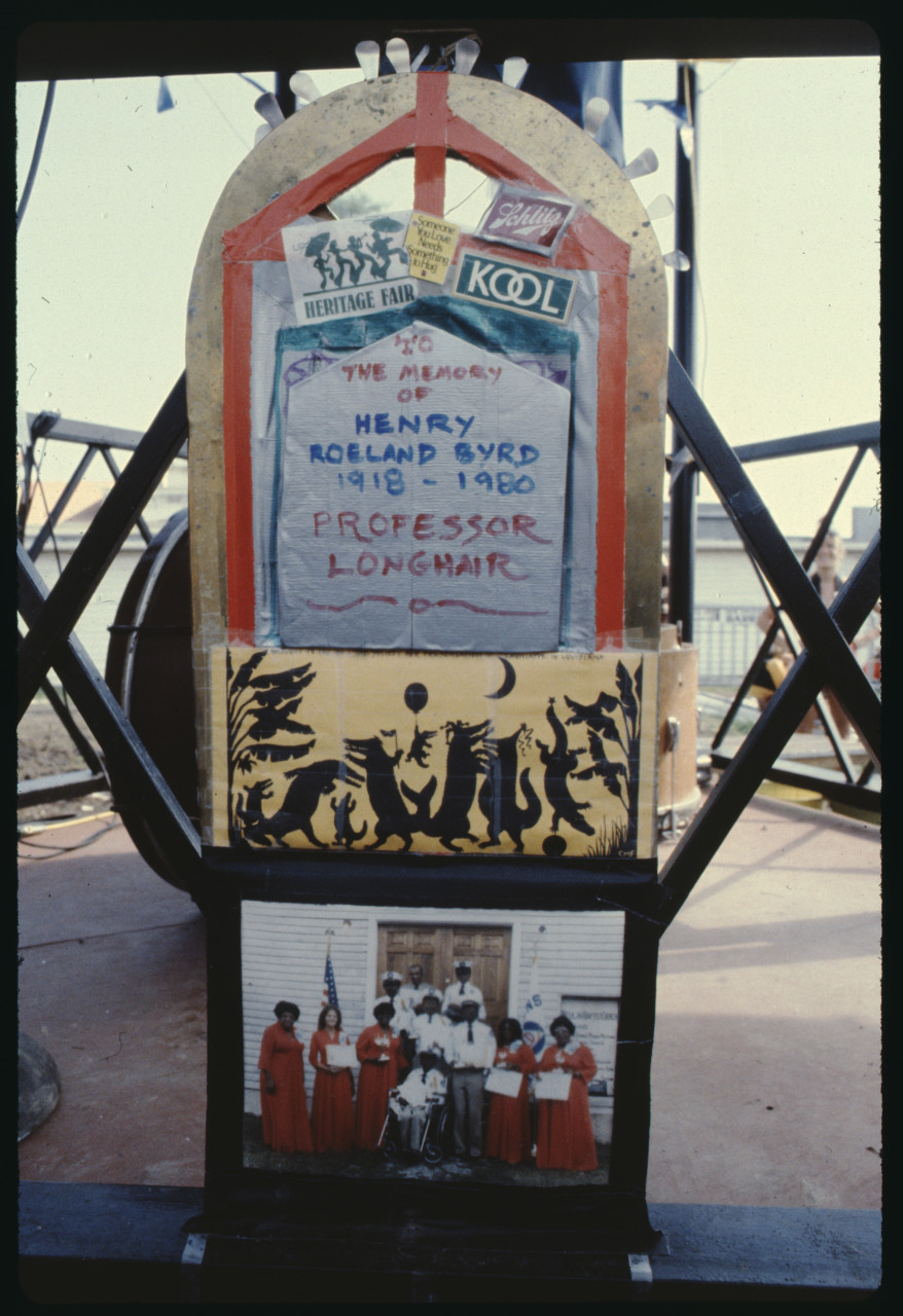 A decorated memorial for Henry Roeland Byrd, also known as Professor Longhair, featuring photos, a sign listing his birth and death years (1918-1980), and various logos and images, including a group of people in red robes holding awards.