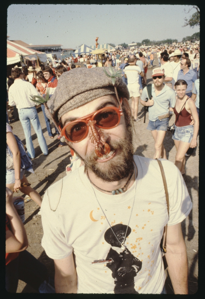 A bearded man with a butterfly on his nose stands in a crowded outdoor festival. Hes wearing red sunglasses, a hat, and a graphic t-shirt. People and tents are visible in the background.