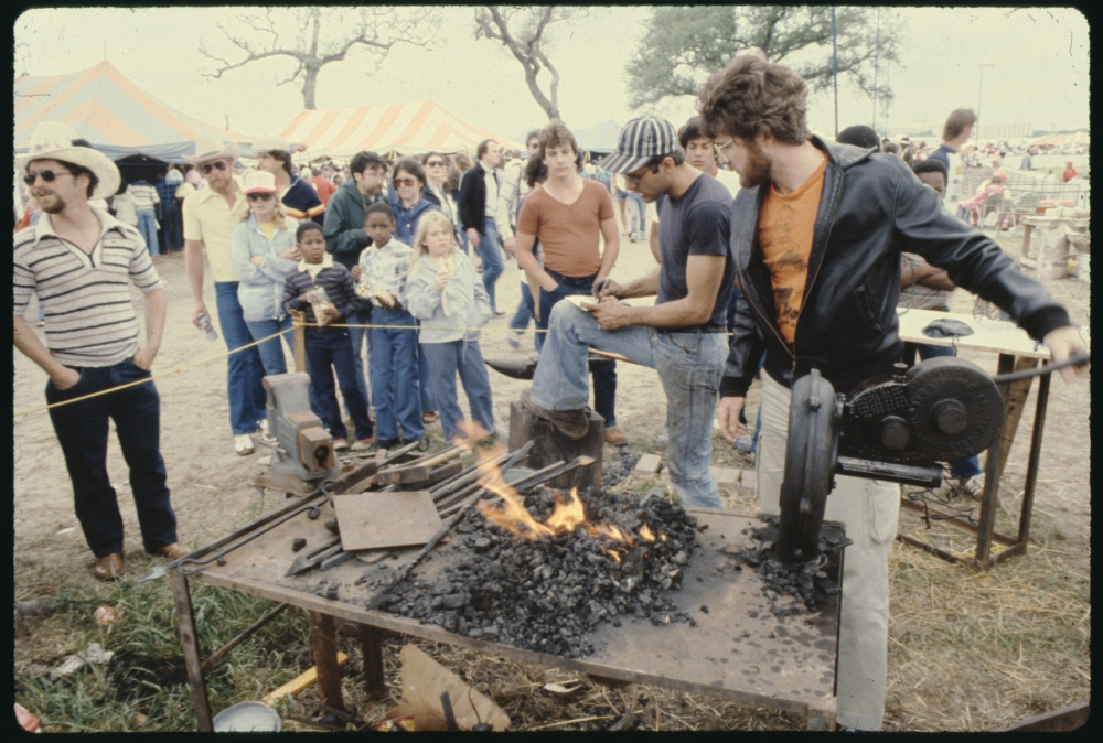A group of people gather around a blacksmith demonstration at an outdoor event. Adults and children watch as two men work with tools near a small forge, with flames visible. Tents and more onlookers are in the background.
