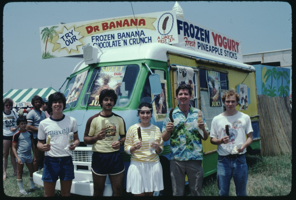 A group of five people standing in front of a colorful ice cream truck labeled Dr. Banana. They are holding frozen treats, with a sign advertising frozen banana and yogurt options above. A line of people is visible to the left.