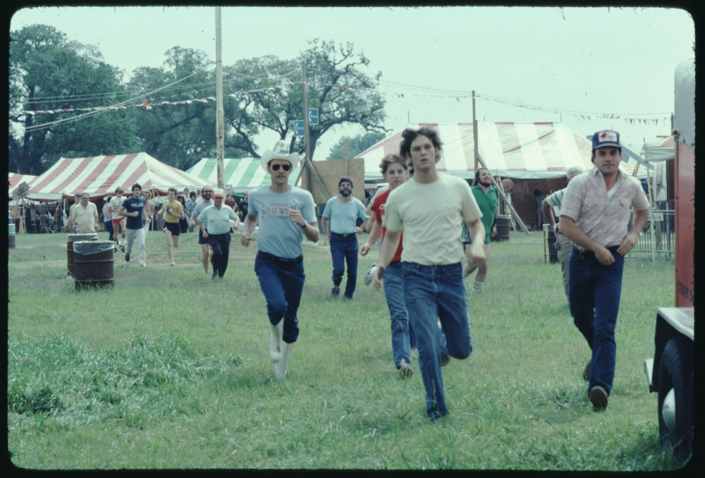 A group of men are running across a grassy field at an outdoor event. Tents with red and white stripes are visible in the background. Some of the men are wearing casual t-shirts and jeans, and one has a baseball cap. It appears lively and festive.