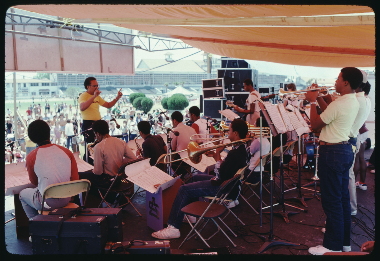 A band performing on an outdoor stage with an audience in the background. The conductor is directing musicians playing trombones, trumpets, and other instruments. The stage is covered by an orange tent, and equipment is visible on the side.