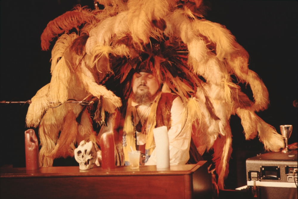 A person sits at a wooden desk, wearing a vibrant headdress made of large brown and orange feathers. The desk is adorned with candles and a skull. The background is dark, highlighting the dramatic attire and props.