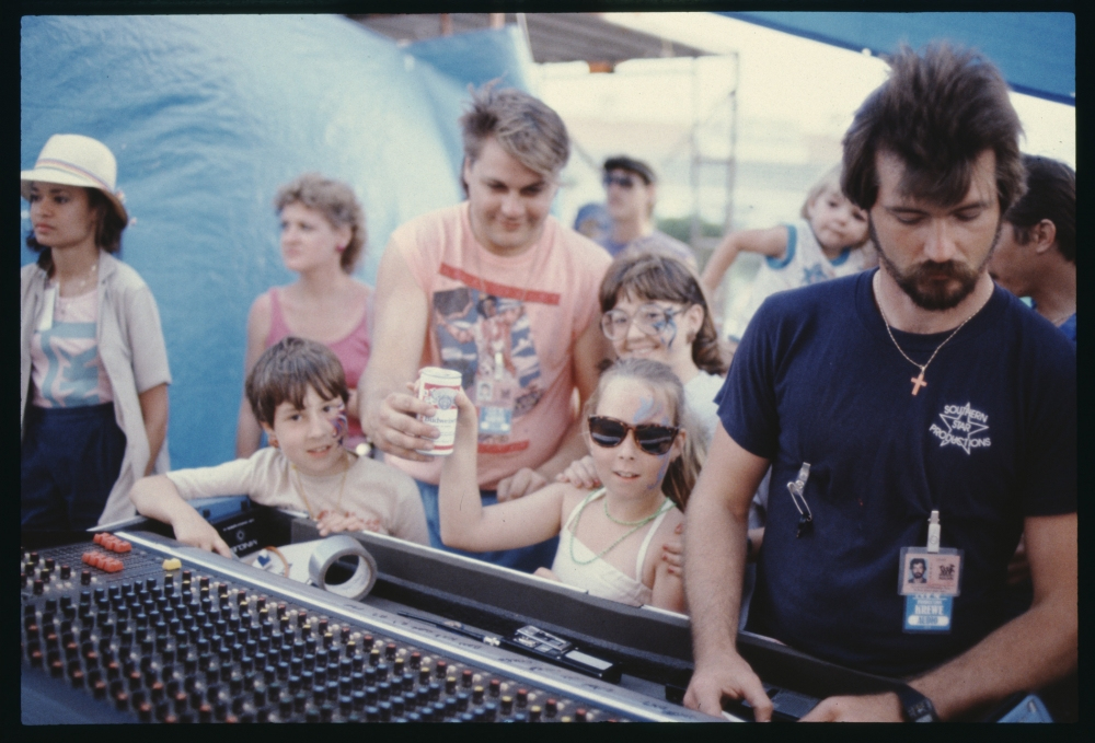 A group of children and adults stand around a sound mixing board at an outdoor event. One child holds a drink can and smiles. A man wearing a navy shirt operates the mixing board. The background features a blue tent and other people.