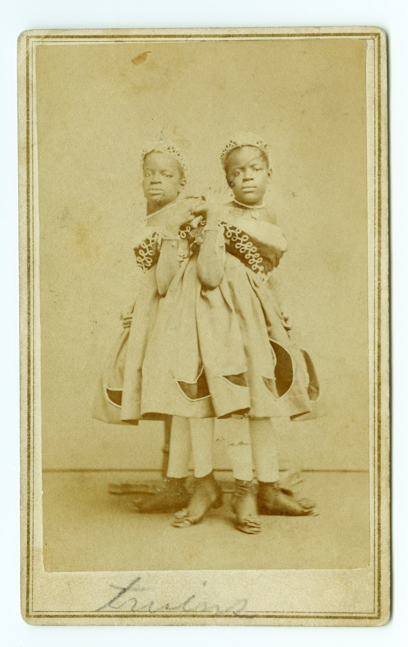 Two young girls are standing side by side, facing the camera. They are wearing identical dresses with decorative designs and matching bows in their hair. The photograph has a sepia tone and is framed with a border.