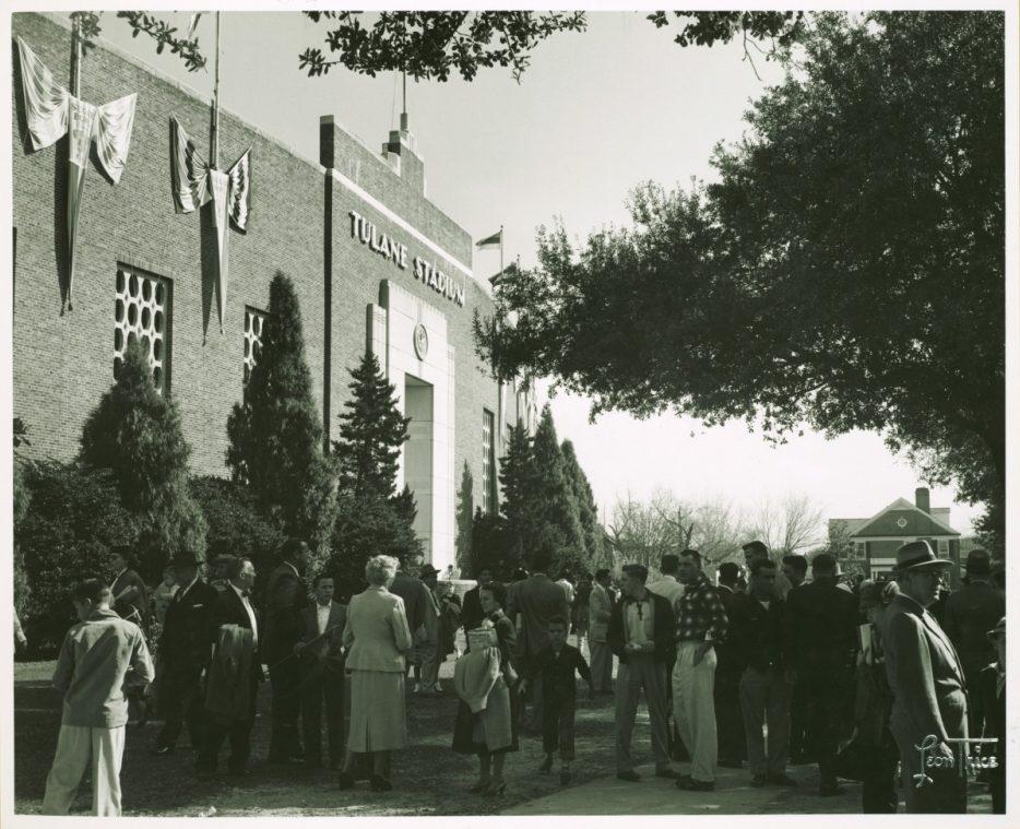 A black and white photo of a crowd gathered outside Tulane Stadium. People are dressed in mid-20th-century clothing, with trees and the stadium entrance visible. Flags adorn the building, and a sunny sky sets a bright scene.