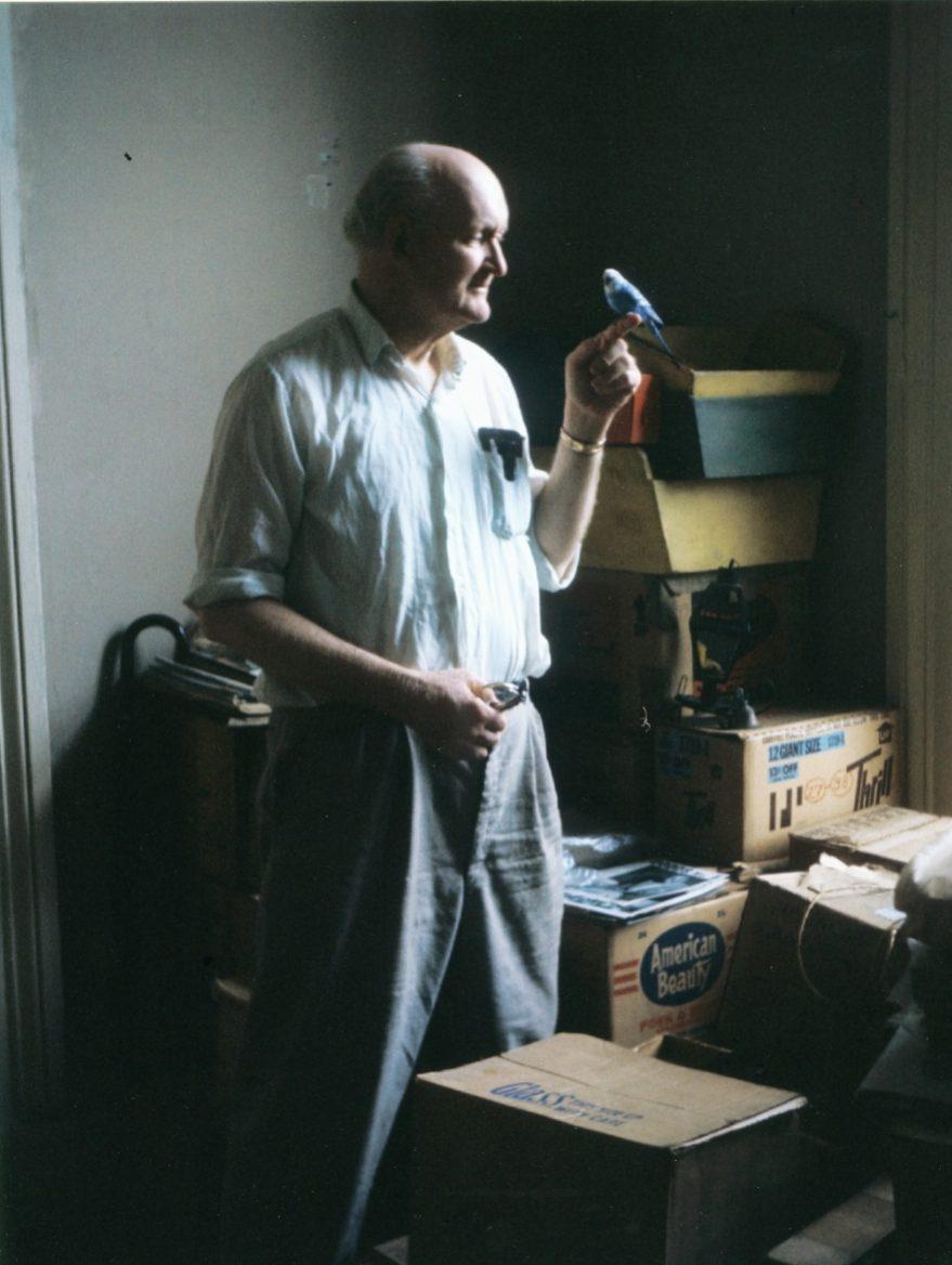 An elderly man stands in a dimly lit room holding a small blue and white bird in one hand. He is wearing a light-colored shirt and dark pants. Cardboard boxes are stacked in the background.