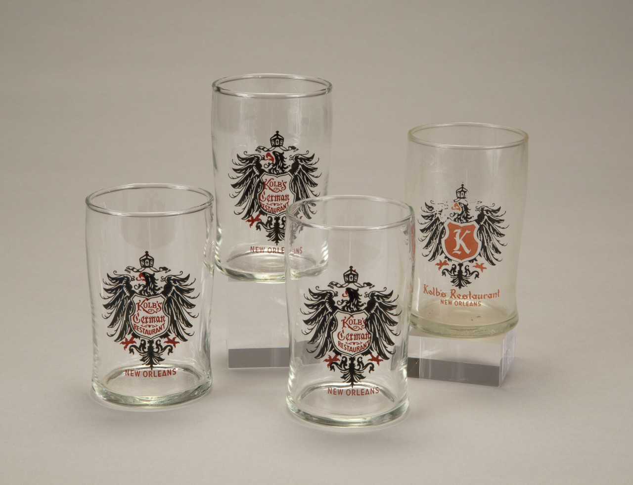 Four clear beer glasses with black and red German-style crest designs labeled Kolbs German Restaurant and New Orleans are arranged on a neutral background.