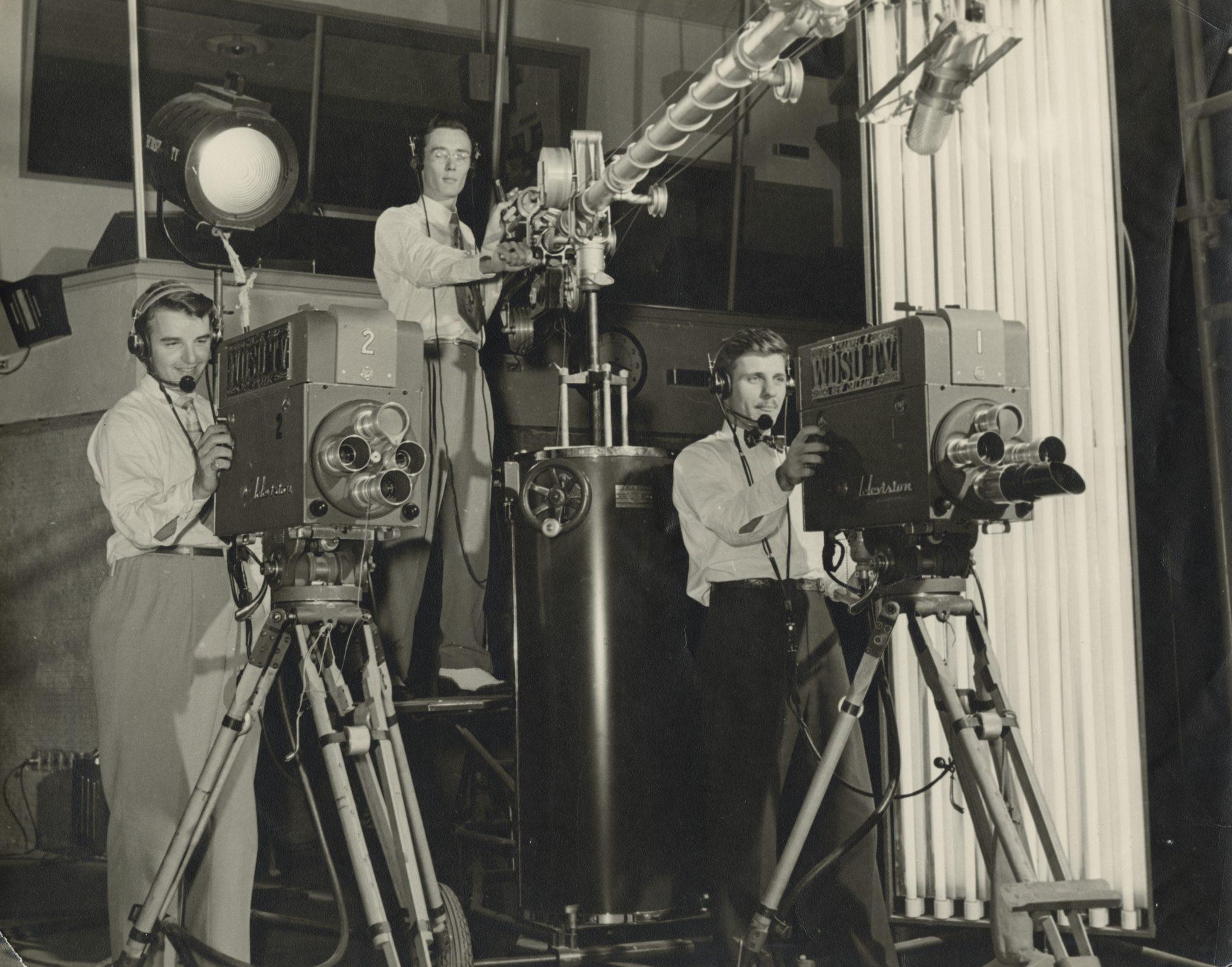 Three men operate vintage television cameras and equipment in a studio. Two men stand behind large film cameras on tripods, while the third adjusts a boom microphone. They wear headphones and classic 1950s attire.