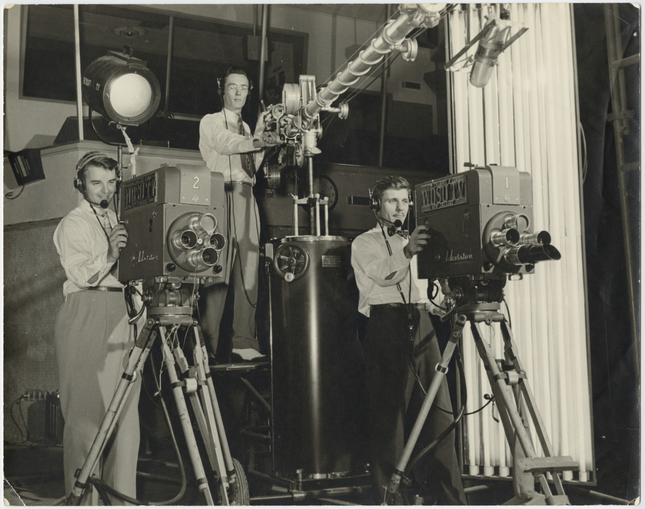 Black and white photo of three men operating vintage film cameras in a studio. Two men are next to cameras on tripods, wearing headsets, while the third adjusts a large camera on a stand. Bright studio lights are visible in the background.