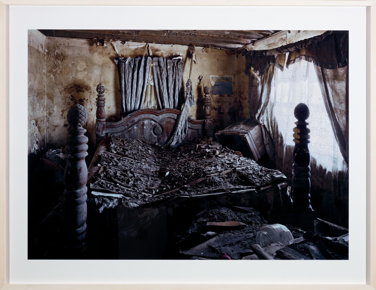 A dilapidated bedroom with a collapsed bed covered in debris. The walls are weathered, and the curtains hang unevenly. Light filters through a lace-covered window, illuminating the rooms disarray.