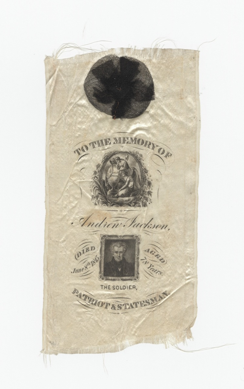 A vintage cloth with a black mourning rosette at the top, featuring a printed tribute to Andrew Jackson. It includes the text To the memory of Andrew Jackson, with his portrait and titles The Soldier, Patriot & Statesman.