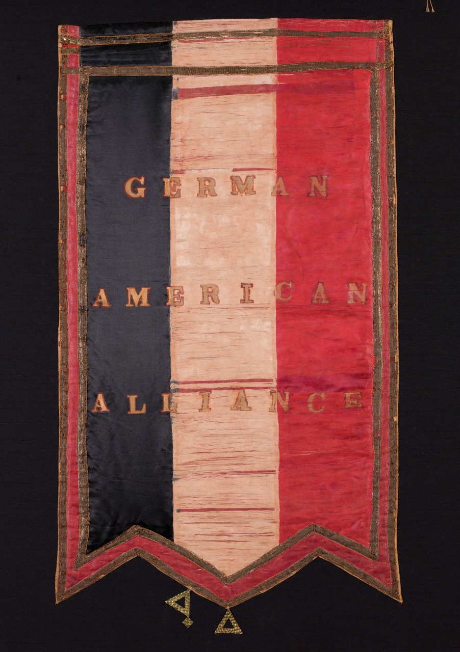 A vertical banner with three colored stripes: black, white, and red. The words German American Alliance are stitched in gold across the stripes. The banner has a decorative triangular bottom edge.