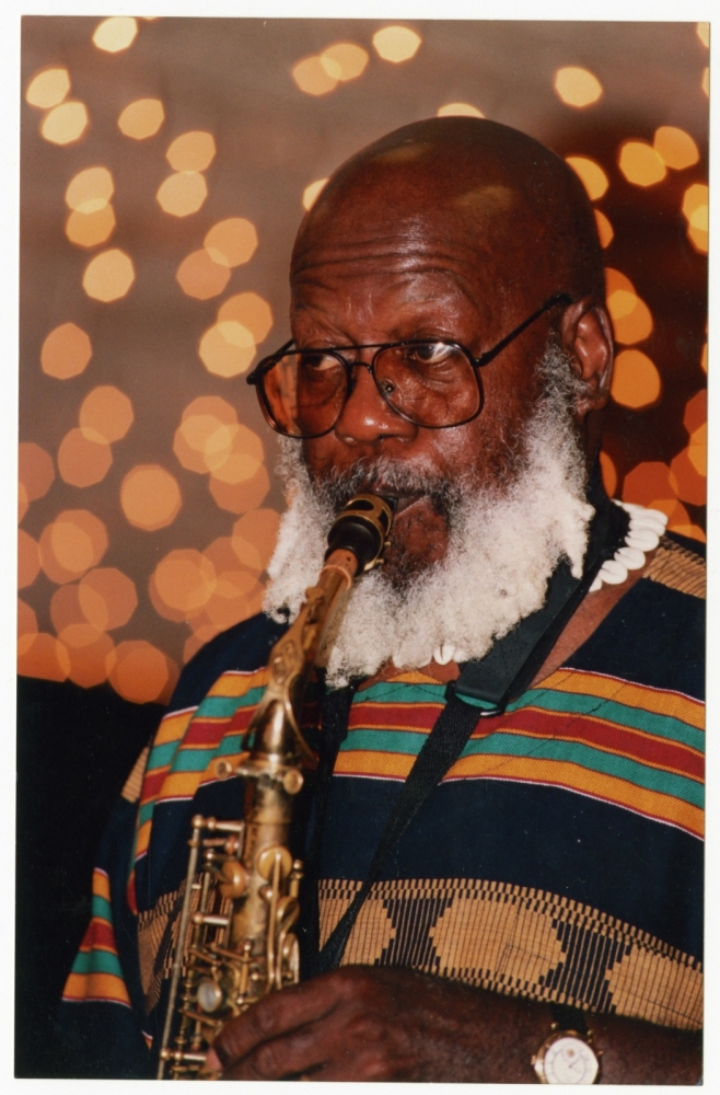 An elderly man with a white beard plays a saxophone, wearing glasses and a colorful, patterned shirt. The background is softly blurred with warm, glowing lights, creating a festive atmosphere.