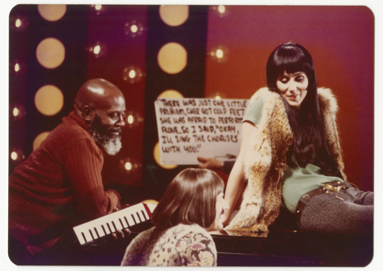 A woman with long dark hair and a fur vest sings into a microphone. A man plays a keyboard while another person sits nearby. The background features stage lights and a sign with lyrics. The setting appears to be a music performance or TV show.