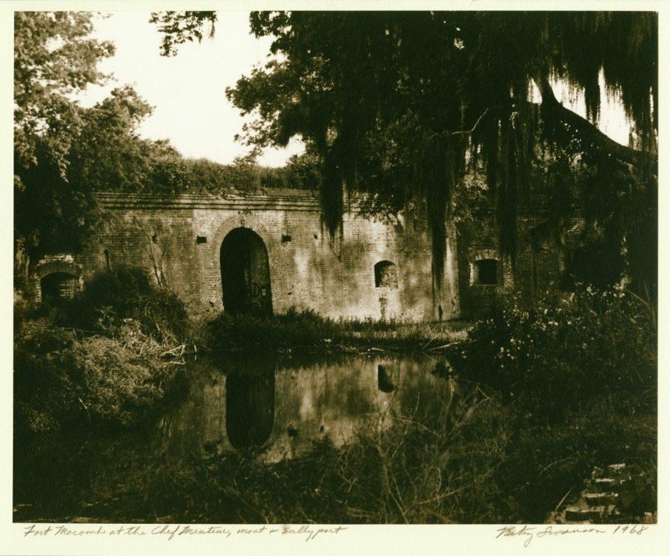 Sepia-toned image of Fort Macomb near a waterway, partially obscured by trees and vegetation. The forts brick archways and walls are reflected in the water. Moss hangs from branches in the foreground, adding to the historic and serene ambiance.