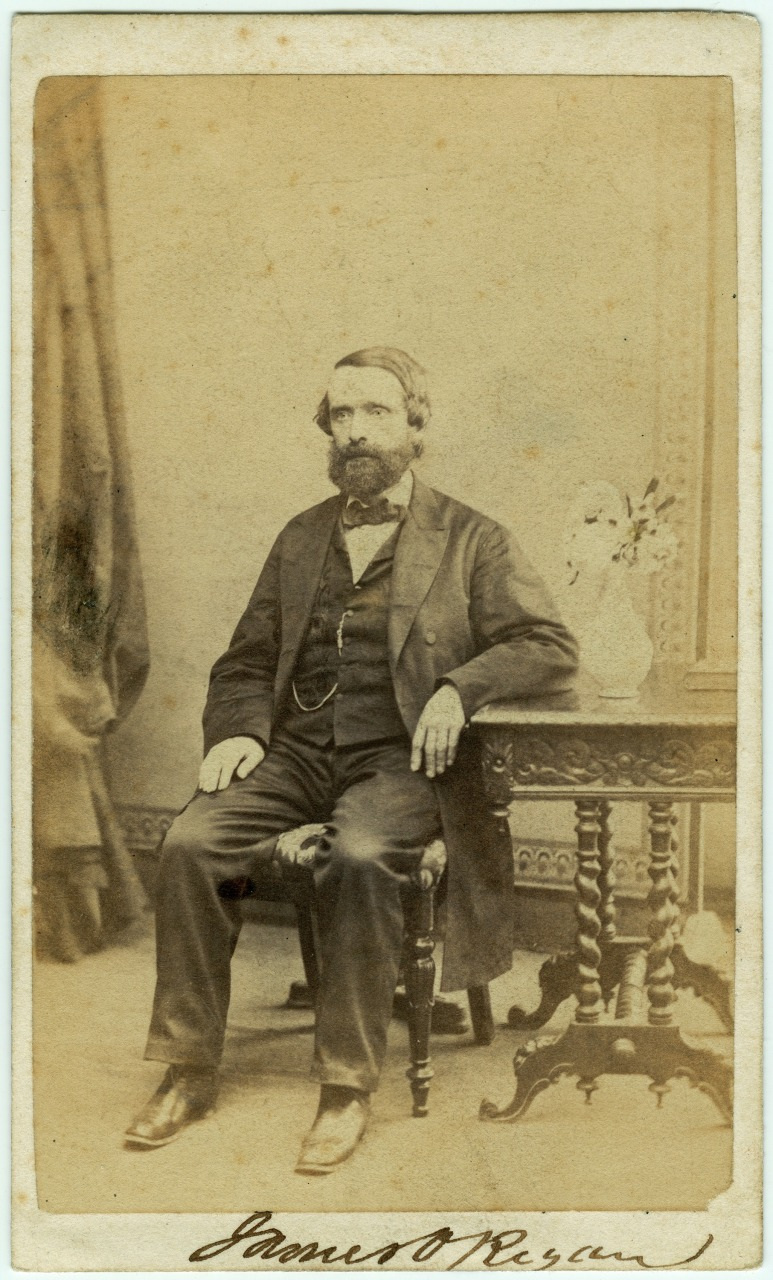 A historical sepia-toned photograph of a seated man in 19th-century attire, resting his arm on an ornate wooden table with a small vase holding flowers. A curtain is partially visible in the background.