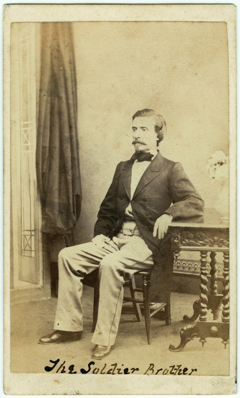 Sepia-toned vintage photograph of a man seated on a chair beside a decorated table, wearing a suit with light pants and a dark jacket. A curtain is partially visible in the background. Text reads The Soldier Brother.