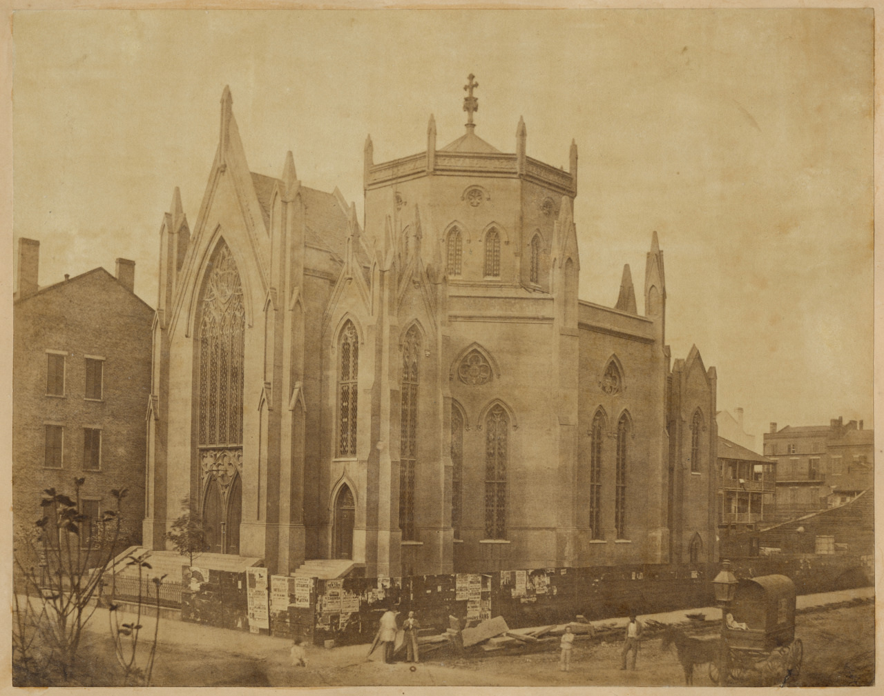 A sepia-toned photograph of a large Gothic-style church with pointed arches, tall spires, and detailed stonework. The building is surrounded by construction materials, and people are visible near a fence in the foreground.