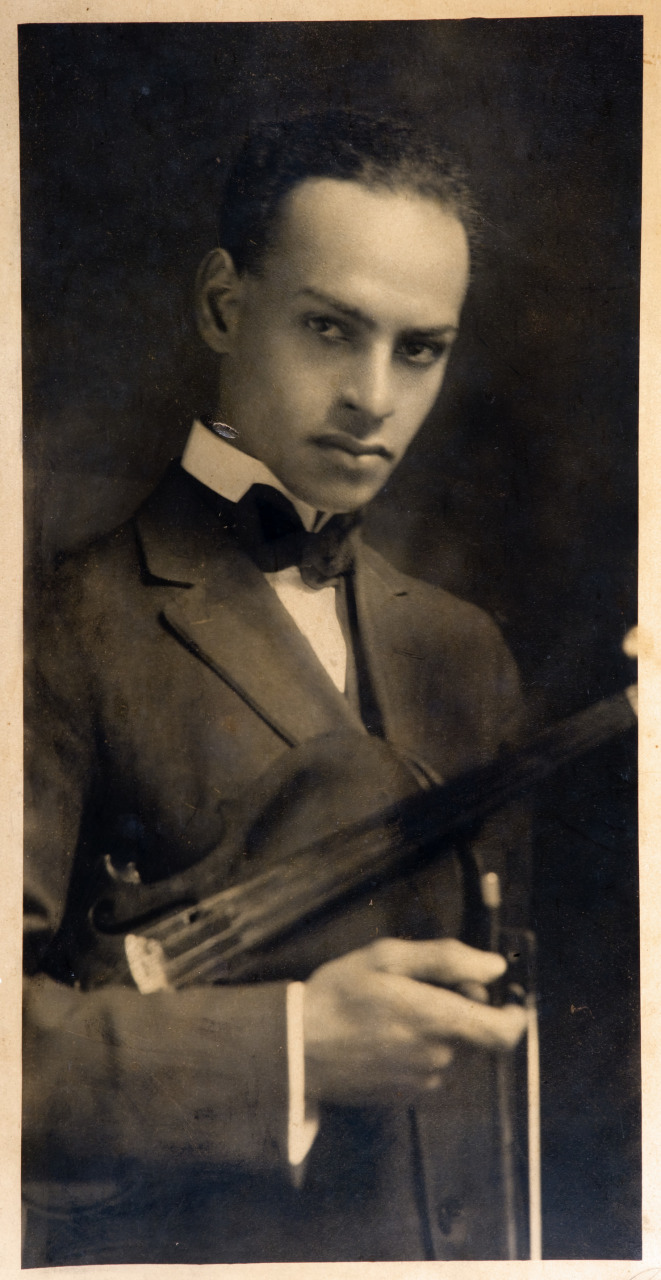 A sepia-toned vintage portrait of a man in a suit holding a violin and bow. He gazes directly at the camera with a serious expression, dressed in a tuxedo with a bow tie. The background is softly blurred.
