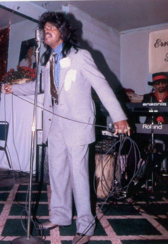 A man in a gray suit sings passionately into a vintage microphone. Behind him, a keyboard player in sunglasses and a hat accompanies him. The scene is set indoors with a banner in the background and a patterned carpet underfoot.
