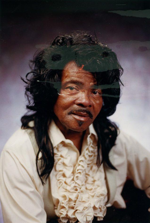 A man with long, dark, wavy hair and a mustache is wearing a cream-colored ruffled shirt. He is seated and looking at the camera against a soft, purple gradient background.