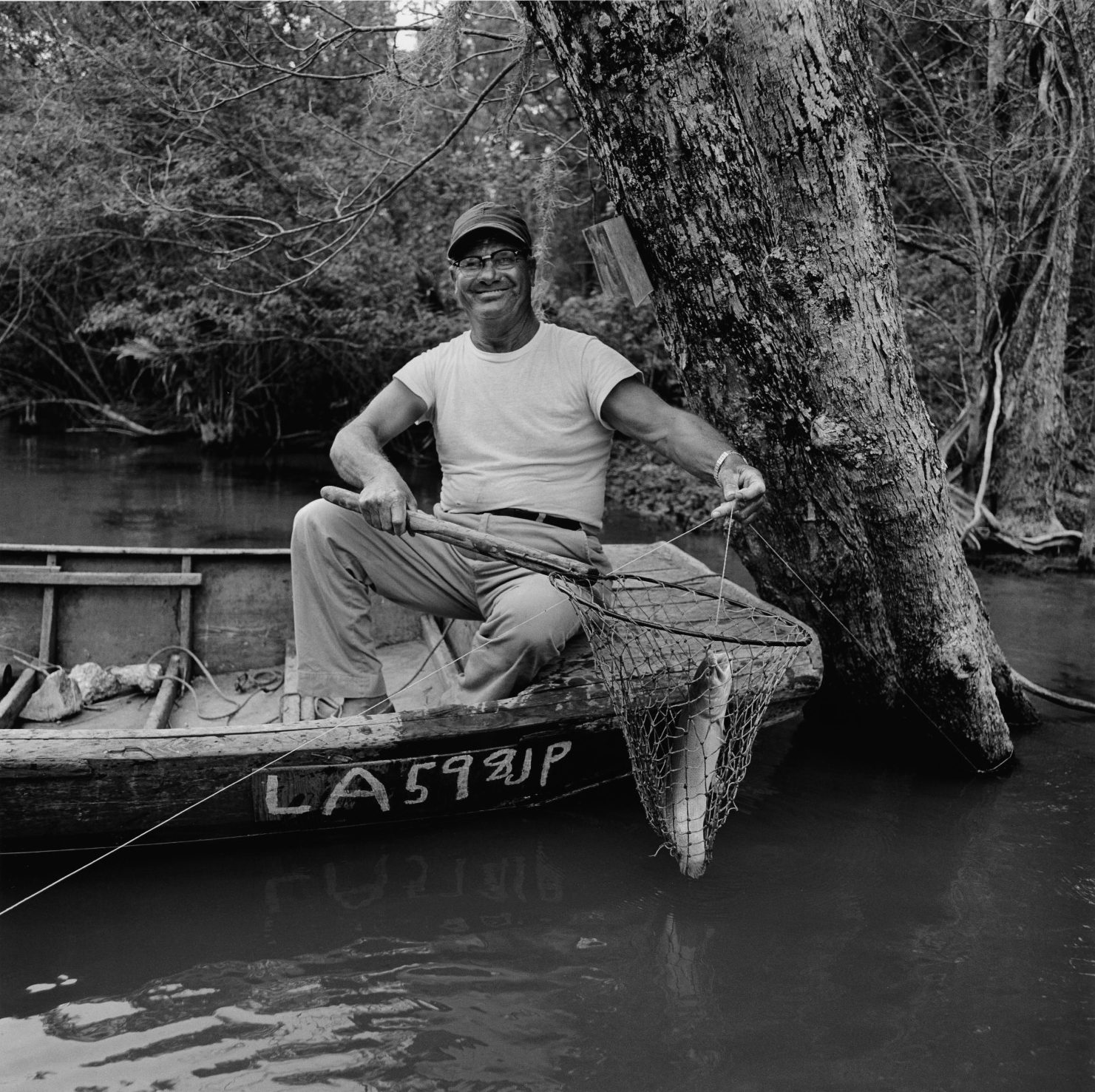 A black and white photo of a fisherman in a small boat with a net, somewhere in Louisiana's marshlands.