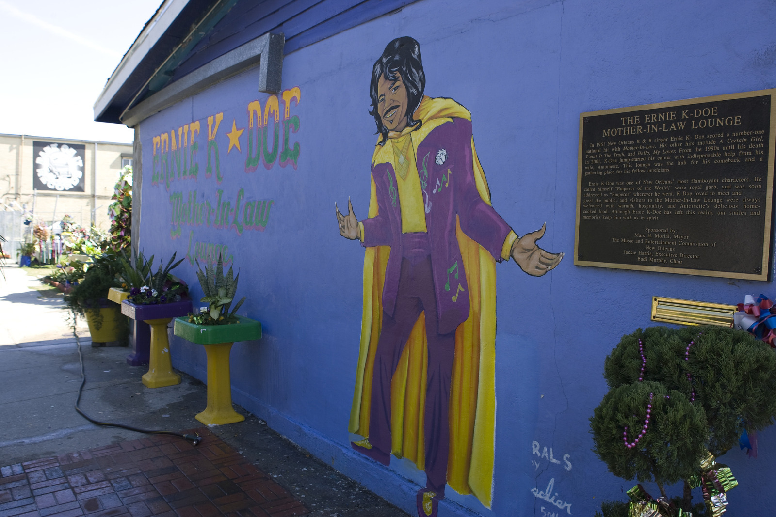 A vibrant mural on a blue wall depicting a person in a purple and yellow outfit. Nearby, a plaque reads The Ernie K-Doe Mother-in-Law Lounge. The scene includes colorful planters and decorations around the building.