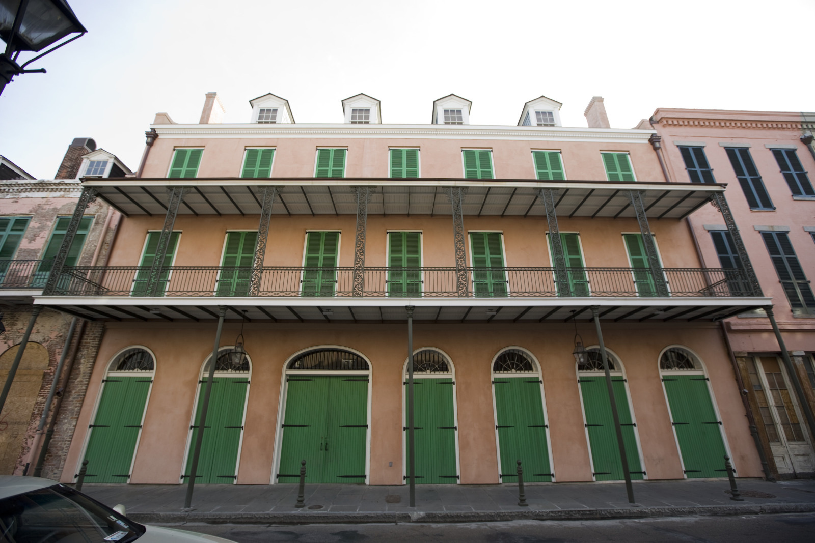 A historic three-story building with a light pink facade and green shutters in New Orleans. The building features green doors, ornate iron balconies, and a row of arched windows on the ground level.