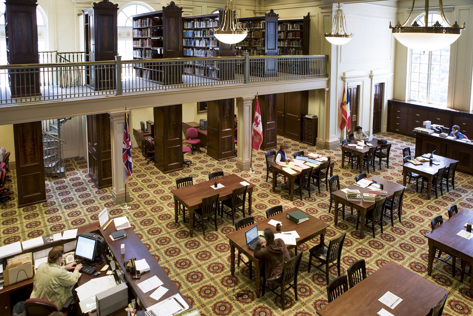 People reading and working at tables in a spacious, well-lit library with high ceilings. Theres a mezzanine level with bookshelves and more seating. Large windows, elegant chandeliers, and a patterned carpet add to the classic ambiance.