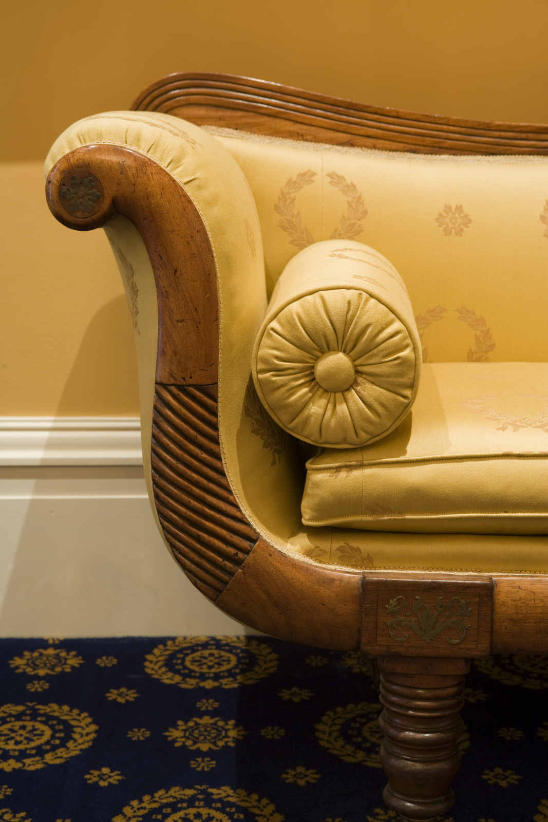 Close-up of an elegant, vintage-style sofa with golden upholstery and intricate woodwork on the armrest. The arm features a round bolster pillow, and the sofa sits on a dark carpet with a gold floral pattern.