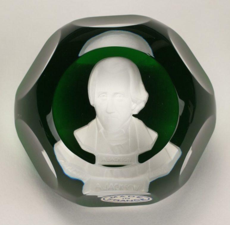 A green and clear glass paperweight featuring an engraved portrait of a historical figure. The image is highlighted within a faceted circular design. A label at the bottom shows a makers mark.